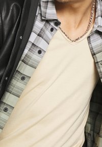 Outfit a strati composto da una t-shirt beige a V, una camicia a quadri grigio e nera con bottoni e una giacca di pelle nera. Dettaglio della catena argento visibile.