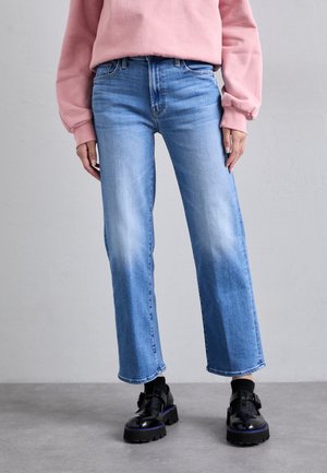 Person iført lyseblå vidde jeans, en pink sweatshirt, sorte sokker og sorte chunky sko med spænde, stående på en grå gulv.