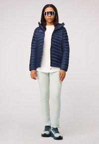 Marine blauwe pufferjack met horizontale stiksels, lichtgroene leggings en blauwe wandelschoenen. Grote spiegelzonnebril maakt de outfit compleet.