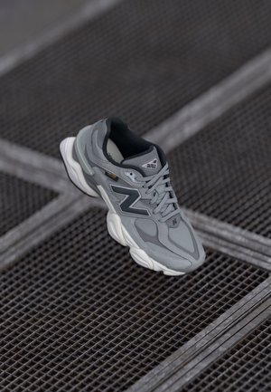 Szaro-biały sneaker New Balance z czarnymi i odblaskowymi detalami na ciemnej metalowej kratce.