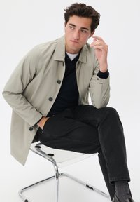 Chaqueta abotonada de color beige claro con una textura ligeramente brillante, llevada sobre una camiseta polo oscura, combinada con jeans negros y zapatos elegantes.