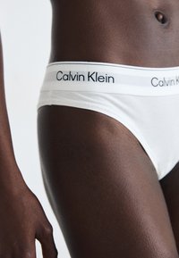 Bijele pamučne gaćice s crnim Calvin Klein logotipom na elastičnom pojasu, glatke teksture, uskog kroja prikazane na nozi.