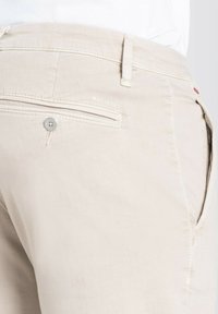 Beige cotton trousers with a single rear pocket, button closure, and embroidered "MACJEANS" logo. Smooth texture and tailored fit.