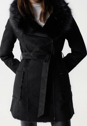 Manteau en suédine noire avec col en fausse fourrure, fermeture éclair, poches avant et ceinture. Texture douce avec une finition lisse.