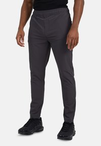 Man in een slim-fit donkergrijze broek en zwarte sneakers, staande tegen een effen witte achtergrond.