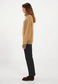 Pull beige oversize avec col roulé côtelé, associé à un pantalon à rayures noires et des chaussures brunes à enfiler. Design simple, texture douce.