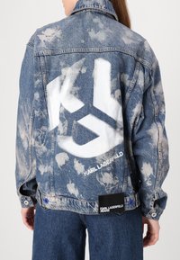 Lys blå denimjakke med slitt finish og hvit logo på ryggen. Har klassisk krage og knappefeste.