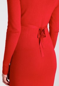 Langärmliges rotes Kleid mit gerippter Textur, das eine taillierte Silhouette und ein gebundenes Taillenakzent aufweist.