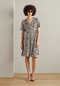 Robe portefeuille en imprimé léopard dans un tissu crème et noir, à manches courtes, avec une jupe évasée, associée à des sandales à lanières beiges.