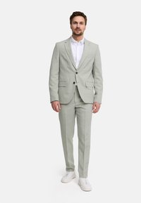Man standing wearing a light gray suit with a white shirt and white sneakers against a plain white background.