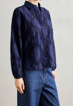 Persoon draagt een marineblauwe blouse met lange mouwen, getextureerde golfpatronen en kraag, gecombineerd met hoge taille blauwe denim jeans.