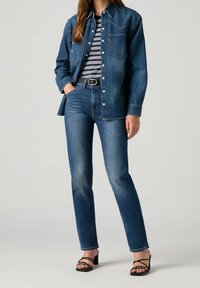 Chemise en denim bleu foncé, avec un bouton à l'avant, deux poches poitrine et un col classique, accompagnée d'un jean en denim de teinte coordonnée. Escarpins noirs à lanières.