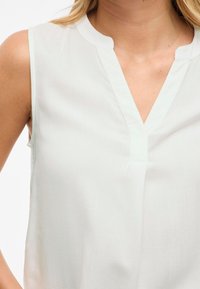 Blusa sin mangas ligera en suave tejido blanco, con escote en V y sutiles detalles de costura. Textura suave, corte holgado.