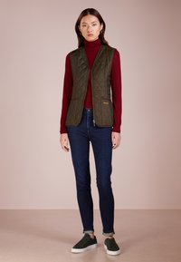 Quilted olivgrüne Weste mit hohem Kragen, kombiniert mit einem figurbetonten burgunderfarbenen Pullover und dunkelblauen Jeans, ergänzt durch dunkelgrüne Sneakers.