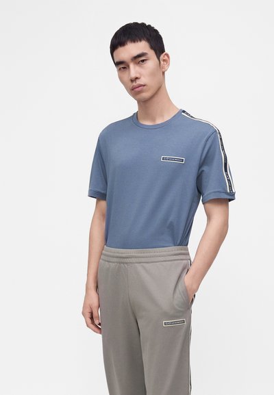 Млад мъж, носещ синя тениска EA7 Emporio Armani и сиви панталони с логота и брандирани ленти на раменете и панталоните.