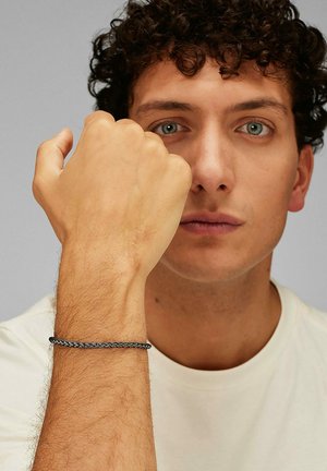 Giovane con capelli ricci indossa una camicia bianca e mostra il pugno chiuso con un braccialetto intrecciato scuro al polso contro uno sfondo grigio.