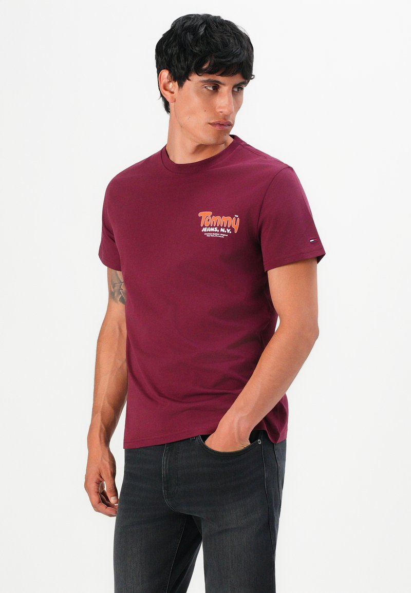 Man in maroon Tommy Jeans T-shirt and dark jeans, standing with one hand in pocket, looking to the side against a white background.