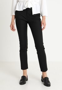 Vrouw in strakke zwarte broek, witte blouse met geknoopte taille, en zwarte loafers, staand tegen een effen lichte achtergrond.