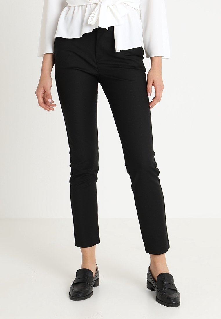 Vrouw in strakke zwarte broek, witte blouse met geknoopte taille, en zwarte loafers, staand tegen een effen lichte achtergrond.