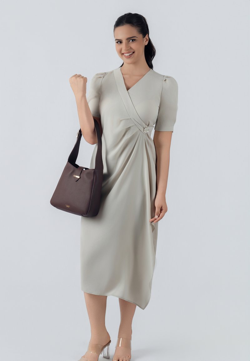 Vestito beige chiaro con un design avvolgente, maniche a sbuffo corte e un anello decorativo. Borsa a tracolla marrone scuro con una texture liscia.