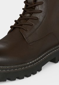 Σκούρο καφέ δερμάτινο μποτάκι με στρογγυλεμένη μύτη, δέσιμο μπροστά, λεπτομερείς ραφές και παχύ μαύρο πέλμα με λευκό διακοσμητικό ρέλι ραφής.