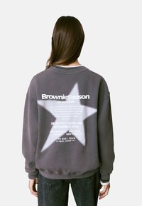 Sweatshirt gris avec un grand motif d'étoile blanche au dos, présentant les détails de l'événement en texte gras. Poignets et ourlet côtelés.