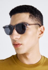 Graue, runde Sonnenbrillen mit einem transparenten Rahmen, ausgestattet mit dunklen Gläsern und dünnen, eleganten Bügeln. Getragen von einer Person mit kurz gewelltem Haar.