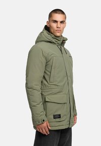 Khaki parka jakk, millel on kapuuts, ette tõmmatav lukk, mitu taskut ja silt all paremas nurgas. Tekstuuriga kangas ja polsterdatud disain.