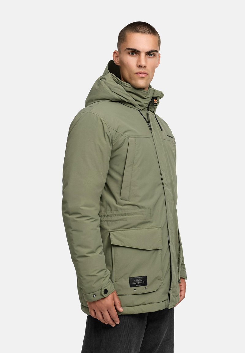 Khaki parka jakk, millel on kapuuts, ette tõmmatav lukk, mitu taskut ja silt all paremas nurgas. Tekstuuriga kangas ja polsterdatud disain.