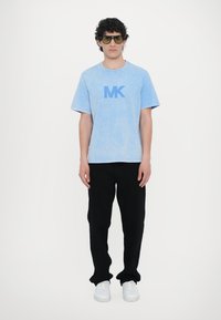 Gaiši zils kokvilnas T-kreklis ar "MK" logo, melni kokvilnas bikses, baltas sporta kurpes. Modelis valkā zaļas saulesbrilles, stāvot pret vienkāršu fona.