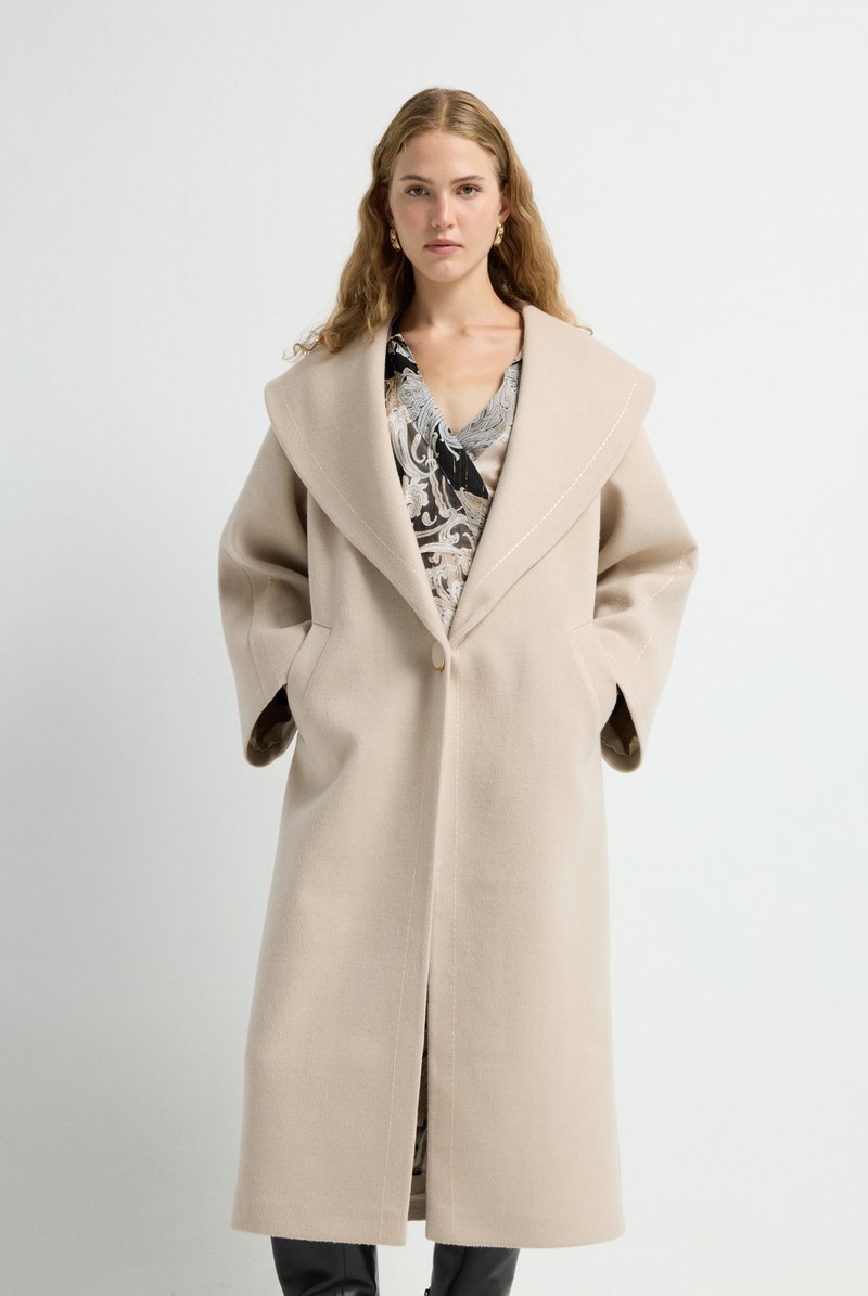 Abrigo de lana beige con cuello grande, cierre frontal con botones, bolsillos grandes y forro en contraste. Llevado sobre una blusa con estampado.