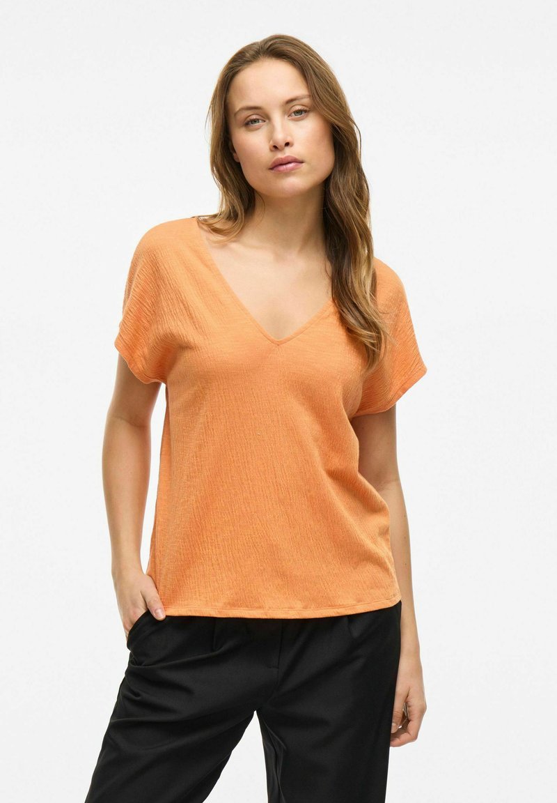 Pomarańczowa bluzka z krótkim rękawem i dekoltem w serek, uszyta z fakturowanej tkaniny. Zestawiona z luźnymi czarnymi spodniami, prezentuje swobodną sylwetkę.