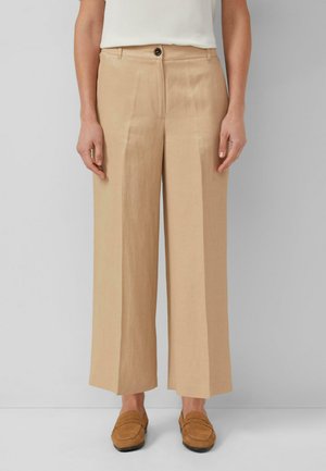Femme portant un pantalon beige à jambes larges avec bouton et fermeture éclair à l'avant, associé à des mocassins en suède marron clair sur un fond gris uni.