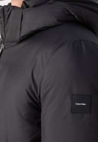 Gros plan d'une personne portant une veste matelassée noire avec un écusson logo Calvin Klein sur le haut du bras.