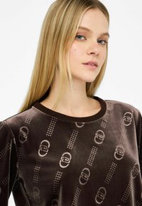 Hnedý velúrový sweatshirt s okrúhlym výstrihom, ktorý obsahuje opakujúci sa vzor loga v svetlejších odtieňoch a štruktúrované akcenty pozdĺž rukávov.