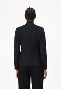 Crni krojeni sako s uskim fitom, reverima i dugmadi na manžetama. Glatka tkanina bez vidljivih uzoraka, uparen s crnim hlačama.