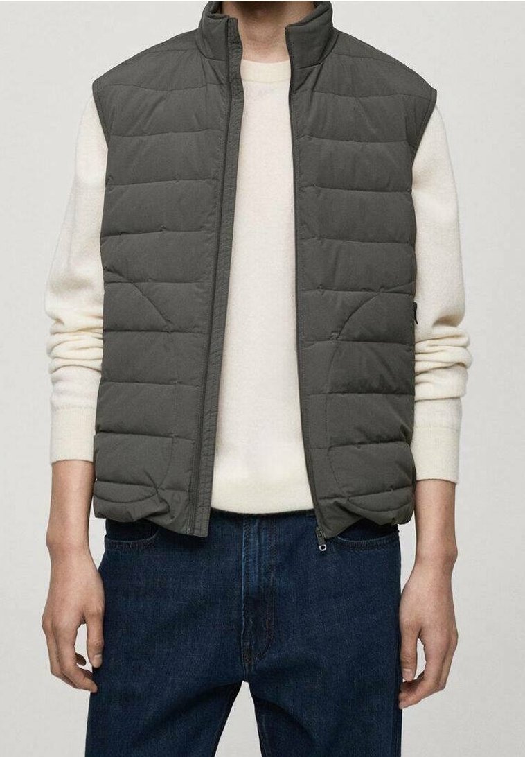 Gilet matelassé en gris foncé, avec une fermeture zippée et un col haut. Il est conçu avec un tissu lisse et des poches latérales, porté sur un pull crème.