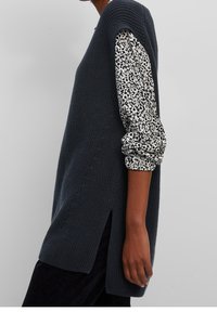 Tunique en tricot gris foncé avec fentes latérales, portée sur une chemise blanche à motifs floraux avec manches bouffantes. Tissu texturé, coupe oversize.