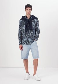 Langarmhemd mit Blättermuster in Blau und Grau, kombiniert mit hellblauen Jeansshorts und weißen Sneakern, mit einem marineblauen Pullover um den Hals gebunden.
