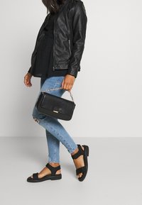 Eine Frau trägt eine schwarze Lederjacke, ein schwarzes Oberteil, ausgefranste hellblaue Jeans, schwarze Sandalen und hält eine schwarze texturierte Handtasche mit goldener Kettenriemen.