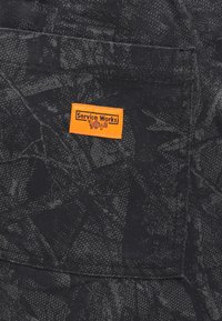 Μαύρο ύφασμα με υφή και αφηρημένο μοτίβο. Μια ορθογώνια πορτοκαλί ετικέτα με την επιγραφή «Service Works» είναι ραμμένη στην επάνω αριστερή τσέπη.