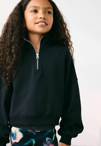Sort zip-sweater med høj krave, løs pasform og ribbet kant. Har en metallisk lynlås og manchetter. Sammen med farverige mønstrede leggings.