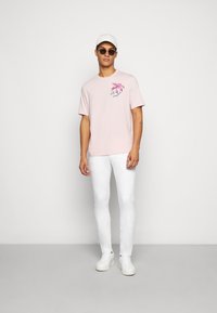Lyserød t-shirt med et frontgrafik af et blomstermotiv og tekst, parret med hvide slim-fit jeans og hvide sneakers. Hvid kasket og solbriller.