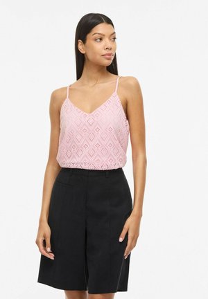 Naine, kes kannab roosat pitsist särkliinikut, millel on õhukesed rihmad, ja musti kõrge vöökohaga laiade säärtega lühkseid pükse, seisab valgel taustal.