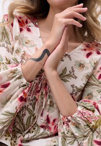 Bluse mit floralem Druck aus weichem Stoff, mit gepufften Ärmeln und elastischen Bündchen sowie einem V-Ausschnitt. Ein Tattoo einer Schlange ist am Handgelenk sichtbar.