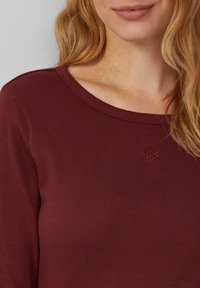 Haut à manches longues bordeaux avec texture côtelée, encolure large et subtil détail de logo brodé. Tissu lisse, coupe ajustée.