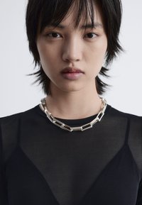 Silberne Kettenhalskette mit rechteckigen Gliedern, getragen über einem transparenten schwarzen Oberteil. Das Oberteil hat eine glatte Textur und eine figurbetonte Passform.