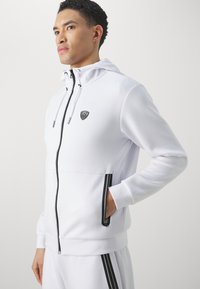 Felpa bianca con zip e cappuccio, con accenti neri, caratterizzata da un patch con logo, cordini regolabili e tasche laterali con zip. Tessuto morbido.
