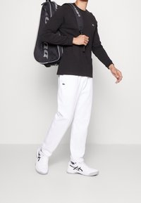 Chemise noire à manches longues, pantalon de survêtement blanc avec logo vert, baskets blanches avec accents noirs, portant un sac de sport noir avec des détails gris.