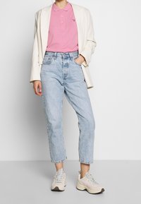 Polo shirt rosa, blazer bianco, jeans a vita alta azzurri chiari e sneakers chiare. Tessuto morbido e vestibilità rilassata con polsini arrotolati.